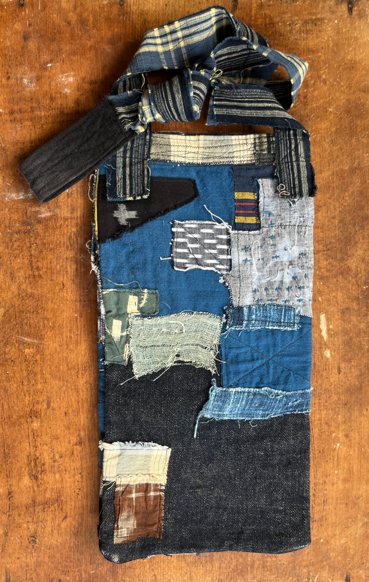 A handmade patchwork fabric bag made from various indigo, blue, black, and beige denim-like fabric pieces, featuring a striped woven strap, photographed flat on a rustic wooden surface.