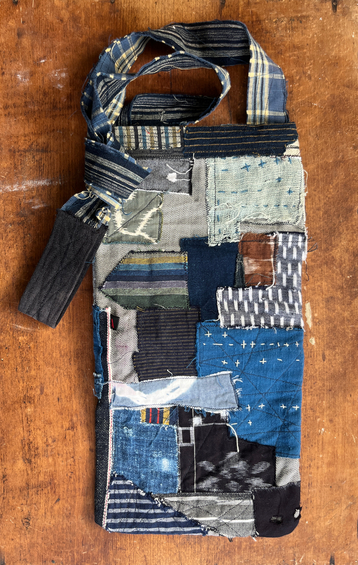 A handmade patchwork fabric bag made from various blue and gray textile scraps, featuring a strap and a matching fabric loop, displayed on a rustic wooden surface.