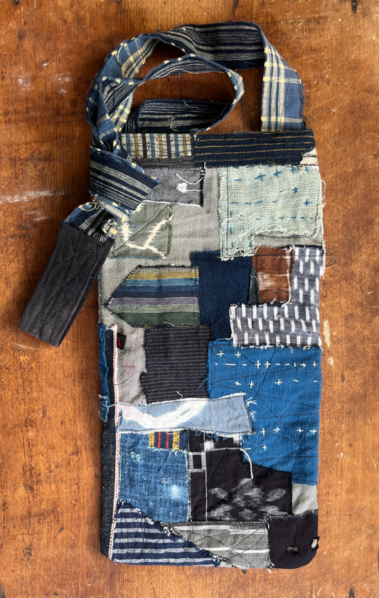 A handmade patchwork fabric bag made from various blue and gray textile scraps, featuring a strap and a matching fabric loop, displayed on a rustic wooden surface.