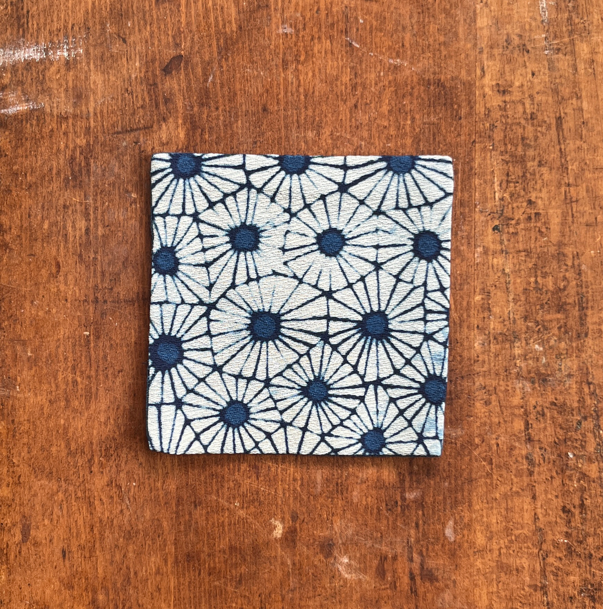 A square fabric coaster or small placemat, featuring a bold indigo flower pattern on a white background, displayed flat on a rustic brown wooden surface.