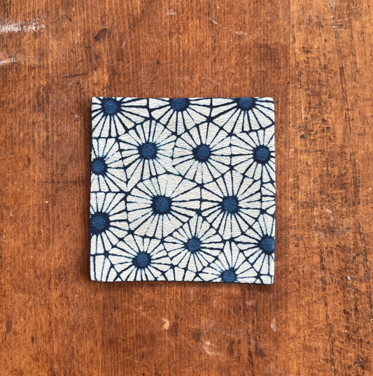 A square fabric coaster or small placemat, featuring a bold indigo flower pattern on a white background, displayed flat on a rustic brown wooden surface.