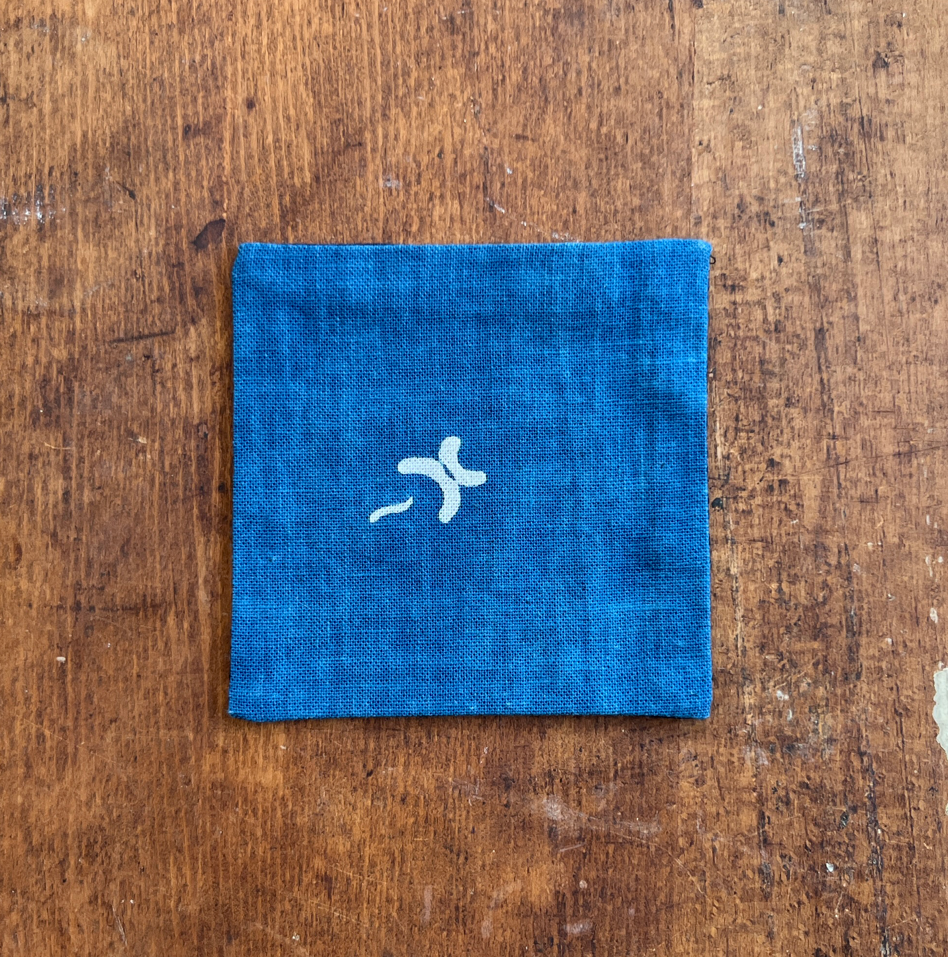 A square fabric coaster or small placemat, featuring a natural motif on an indigo background, displayed flat on a rustic brown wooden surface.