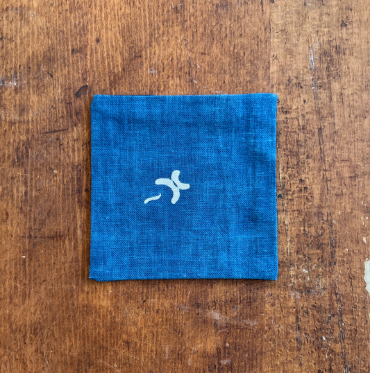 A square fabric coaster or small placemat, featuring a natural motif on an indigo background, displayed flat on a rustic brown wooden surface.