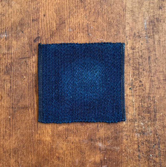 An indigo square fabric coaster or small placemat, displayed flat on a rustic brown wooden surface.
