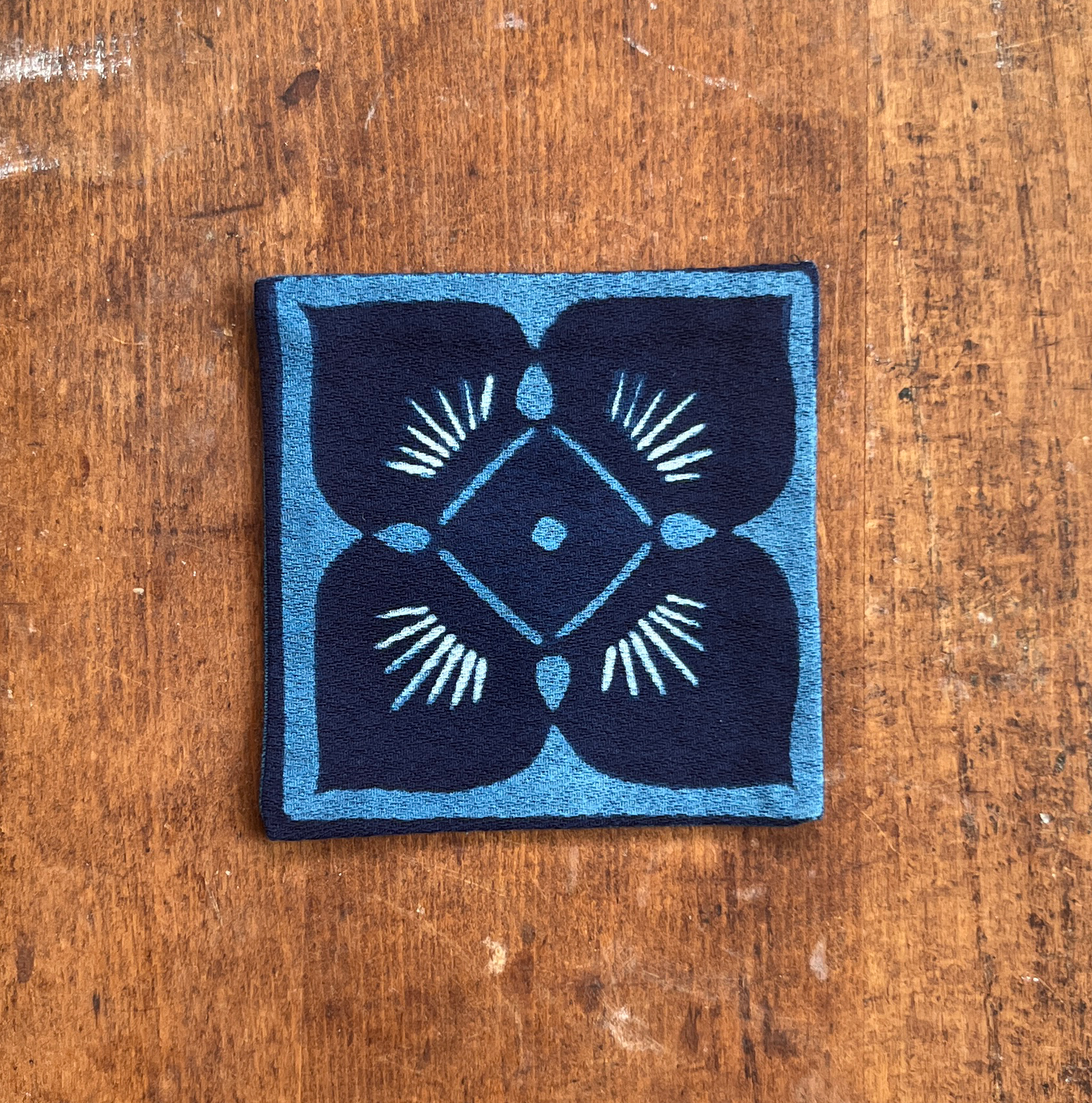 A square fabric coaster or small placemat, featuring a blue flower motif on an indigo background, displayed flat on a rustic brown wooden surface.