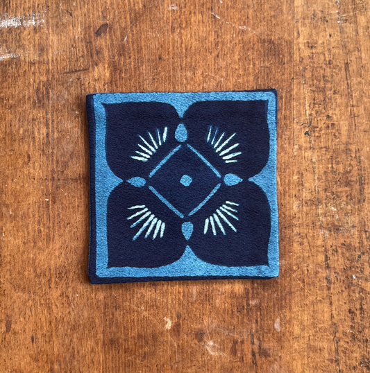 A square fabric coaster or small placemat, featuring a blue flower motif on an indigo background, displayed flat on a rustic brown wooden surface.
