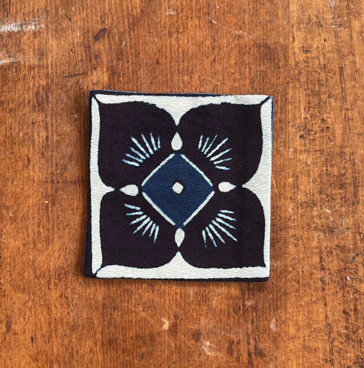 A square fabric coaster or small placemat, featuring a blue flower motif on a white background, displayed flat on a rustic brown wooden surface.