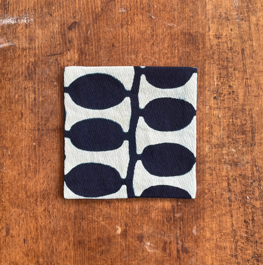 A square fabric coaster or small placemat, featuring a bold indigo leaf pattern on a white background, displayed flat on a rustic brown wooden surface.

