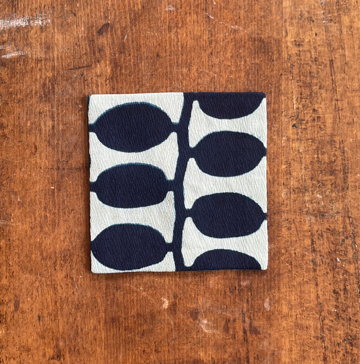 A square fabric coaster or small placemat, featuring a bold indigo leaf pattern on a white background, displayed flat on a rustic brown wooden surface.
