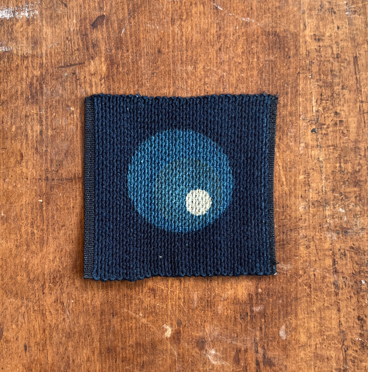 A square fabric coaster or small placemat, featuring a large blue and small white circle motif on an indigo background, displayed flat on a rustic brown wooden surface.
