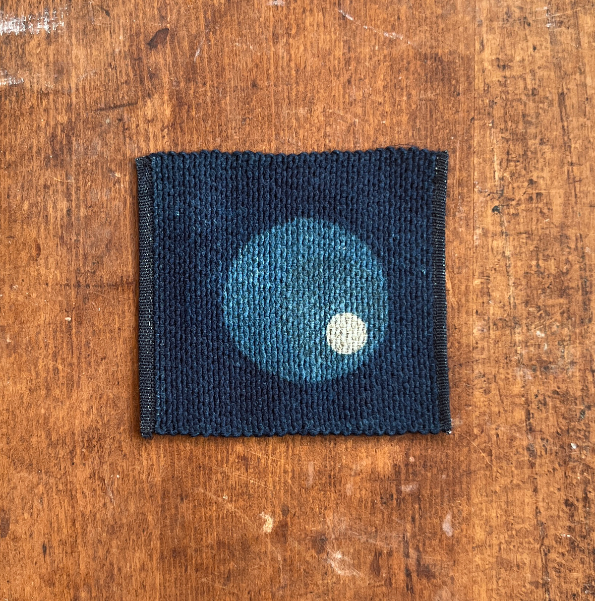A square fabric coaster or small placemat, featuring a large blue and small white circle motif on an indigo background, displayed flat on a rustic brown wooden surface.