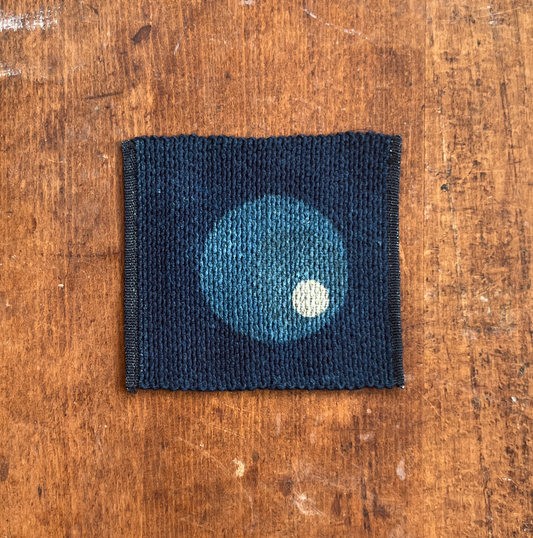 A square fabric coaster or small placemat, featuring a large blue and small white circle motif on an indigo background, displayed flat on a rustic brown wooden surface.