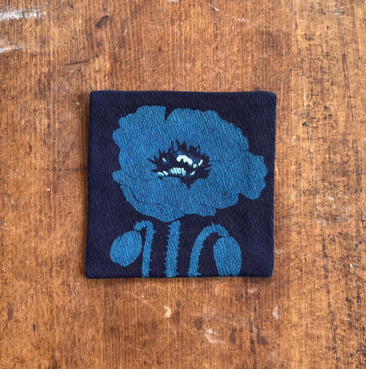 A square fabric coaster or small placemat, featuring a blue flower motif on an indigo background, displayed flat on a rustic brown wooden surface.