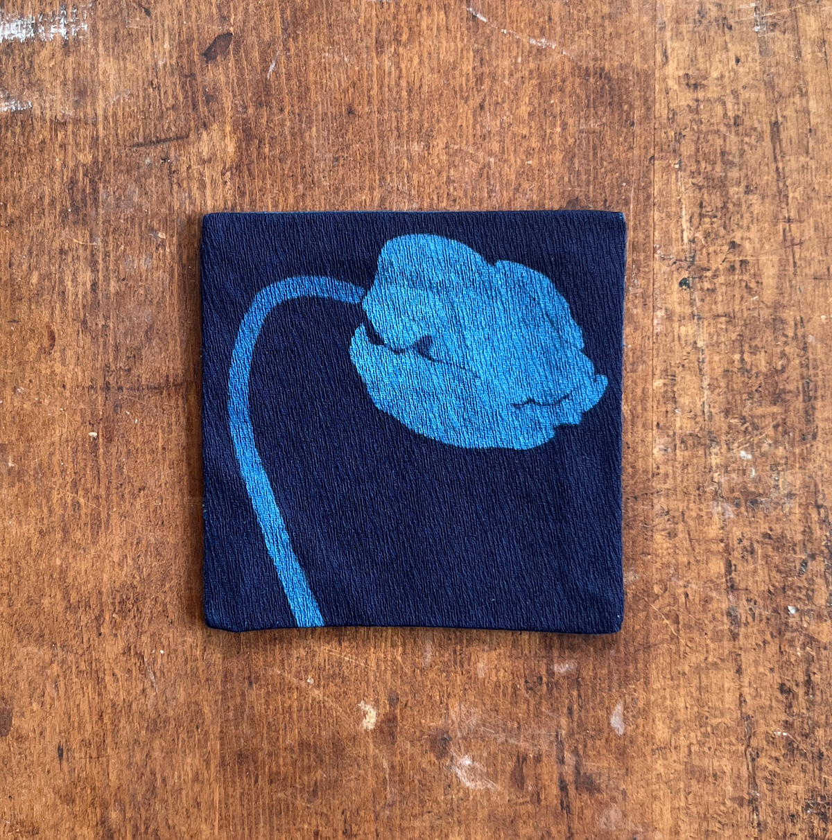 A square fabric coaster or small placemat, featuring a blue flower motif on an indigo background, displayed flat on a rustic brown wooden surface.