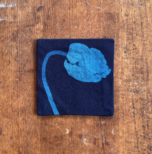 A square fabric coaster or small placemat, featuring a blue flower motif on an indigo background, displayed flat on a rustic brown wooden surface.