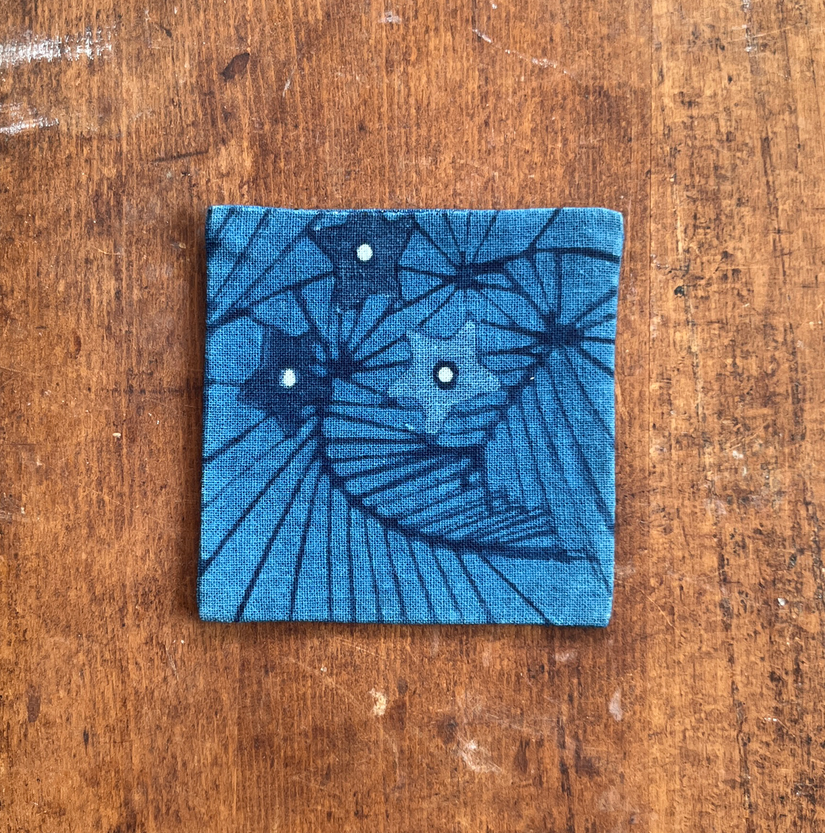A square fabric coaster or small placemat, featuring a starburst and leaf pattern, on an indigo background, displayed flat on a rustic brown wooden surface.
