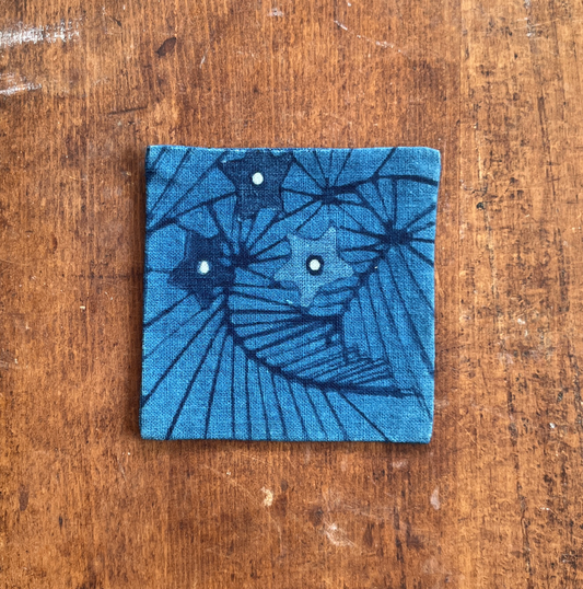 A square fabric coaster or small placemat, featuring a starburst and leaf pattern, on an indigo background, displayed flat on a rustic brown wooden surface.
