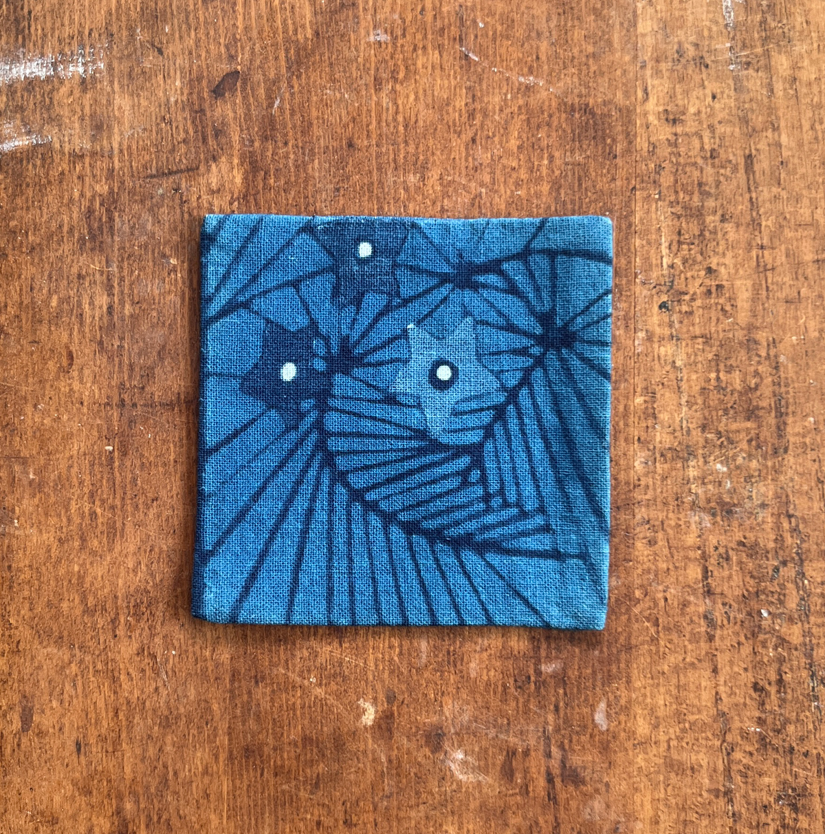 A square fabric coaster or small placemat, featuring a starburst and leaf pattern, on an indigo background, displayed flat on a rustic brown wooden surface.
