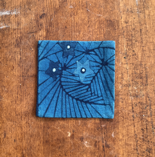A square fabric coaster or small placemat, featuring a starburst and leaf pattern, on an indigo background, displayed flat on a rustic brown wooden surface.
