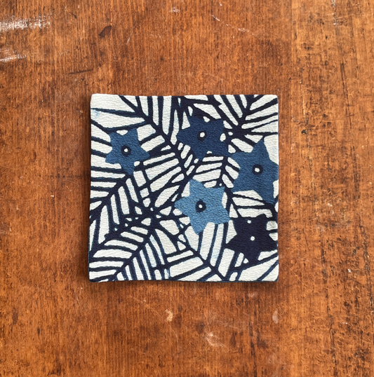 A square fabric coaster or small placemat, featuring a starburst and leaf pattern, on a white background, displayed flat on a rustic brown wooden surface.